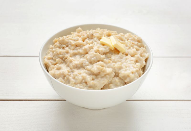 A bowl of creamy oatmeal topped with a pat of butter, served on a white wooden surface.